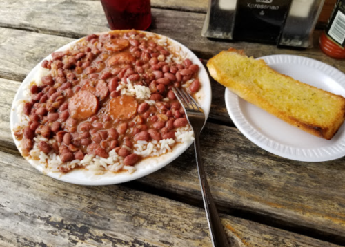 A plate of rice and red beans with sausage, accompanied by a slice of garlic bread, on a wooden table.