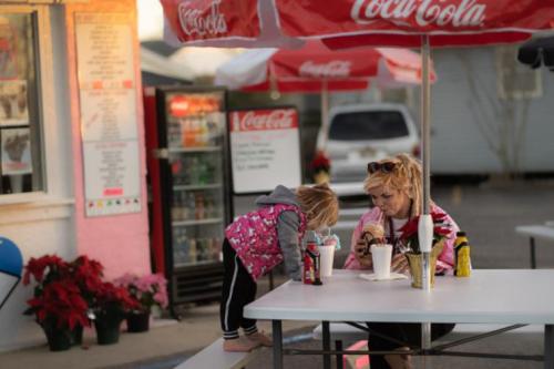 A woman sits at a table with drinks while a child leans over, both enjoying a colorful outdoor setting.