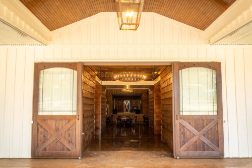 A rustic barn entrance with wooden doors, leading to a warmly lit interior with chandeliers and wooden decor.