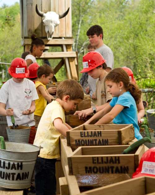 Children and adults work together at a hands-on activity station, wearing hard hats and focused on their tasks.