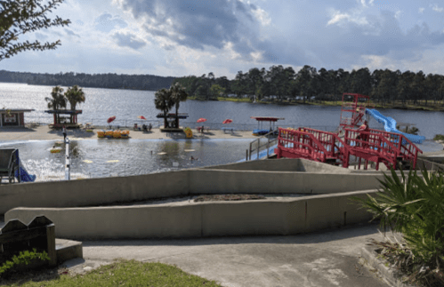A lakeside view featuring a water slide, colorful umbrellas, and recreational areas with kayaks and paddle boats.