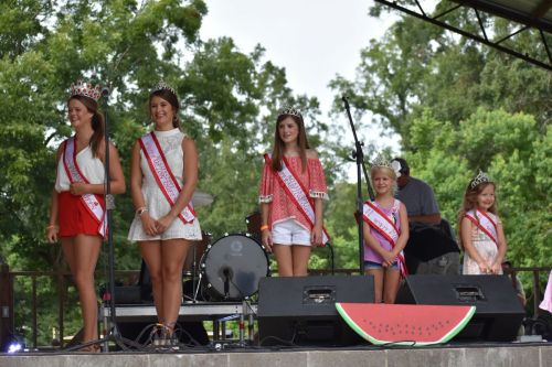 Five young women wearing crowns and sashes stand on stage, smiling at an outdoor event with greenery in the background.