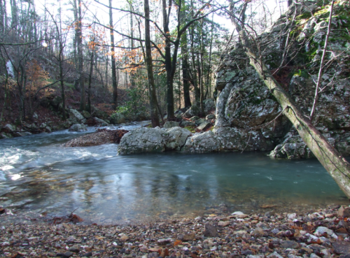 A serene river flows past rocky banks and trees, surrounded by autumn foliage in a peaceful natural setting.