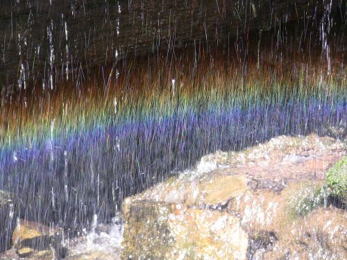A vibrant rainbow forms in mist from a waterfall, with colorful bands shimmering against a rocky backdrop.