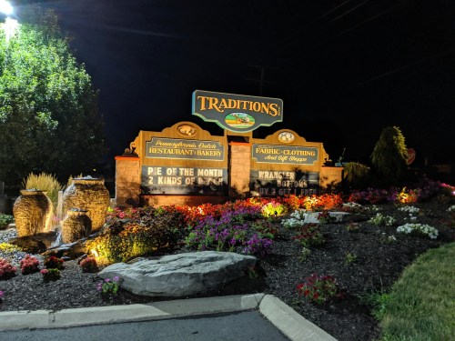 Sign for "Traditions" restaurant and fabric shop, featuring flowers and a water feature, illuminated at night.