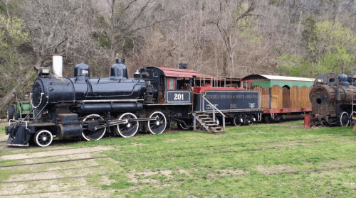 A vintage steam locomotive with a red and black color scheme, parked on grass near trees.