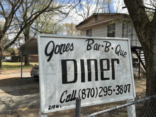 Sign for Jones Bar-B-Que Diner, featuring a phone number and address, set against a backdrop of trees and a building.