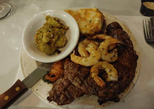 A plate featuring a large steak, grilled shrimp, a side of guacamole, and a cheesy potato dish.