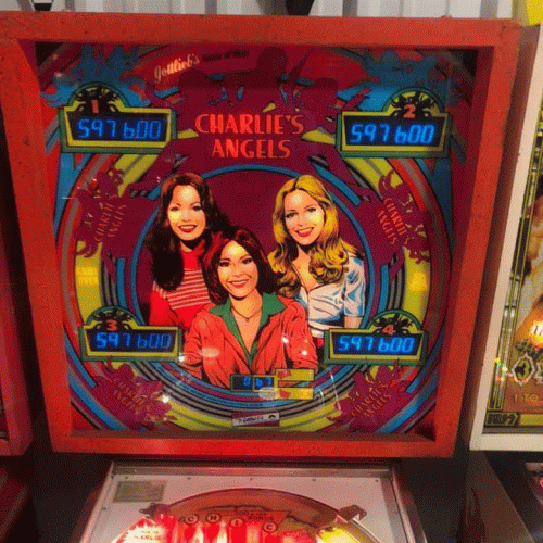 Colorful pinball machine display featuring three women, titled "Charlie's Angels," with scoreboards showing points.