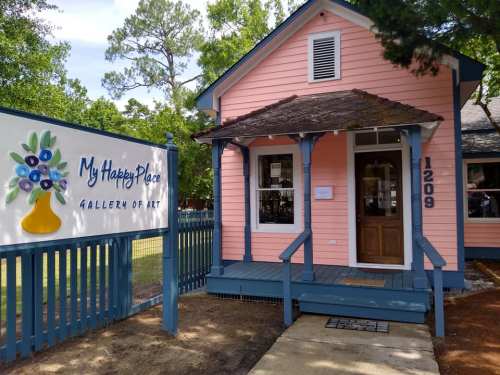 A pink house with a blue porch and sign reading "My Happy Place Gallery of Art" in a green, tree-filled area.