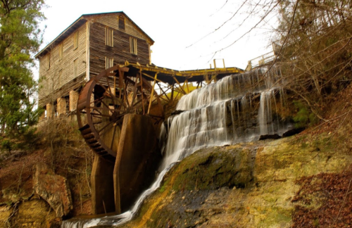 A rustic watermill with a large wooden wheel, cascading water, and surrounded by trees and rocky terrain.
