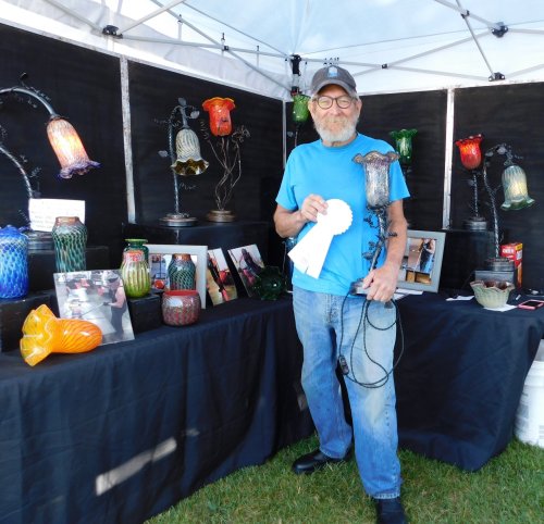 An artist stands in front of a display of colorful glass sculptures, holding a blue ribbon award.