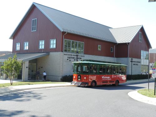 A red trolley bus parked in front of a modern visitors center with a metal roof and large windows.