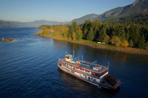 A scenic view of a riverboat cruising on a calm river, surrounded by lush green trees and mountains in the background.