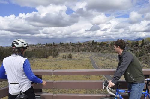 Two cyclists on a bridge overlook a scenic landscape with rolling hills and a winding path under a cloudy sky.