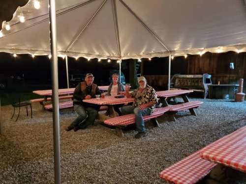 Three people sitting at picnic tables under a tent, enjoying drinks in a cozy outdoor setting with string lights.