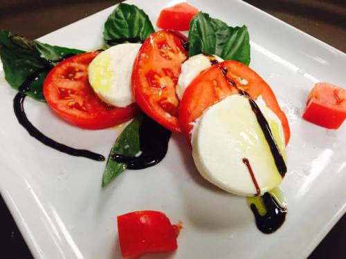 A fresh Caprese salad with sliced tomatoes, mozzarella, basil, and drizzled balsamic glaze on a white plate.