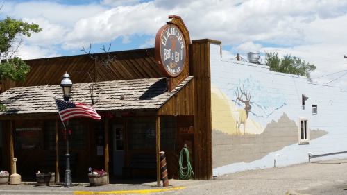 A rustic bar and grill with a wooden exterior, featuring a mural of an elk and an American flag out front.