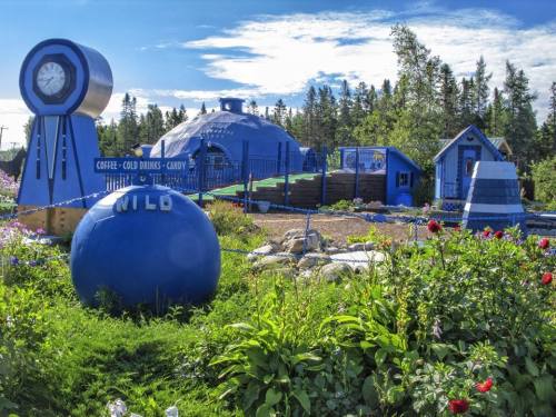 A whimsical blue building with a clock, surrounded by colorful flowers and trees, featuring play areas and signage.