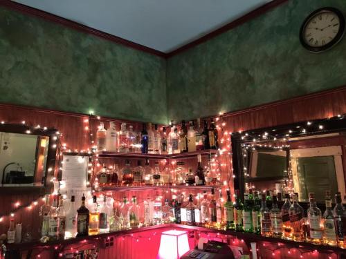 A cozy bar corner with shelves of various liquor bottles, adorned with twinkling lights and a clock on the wall.