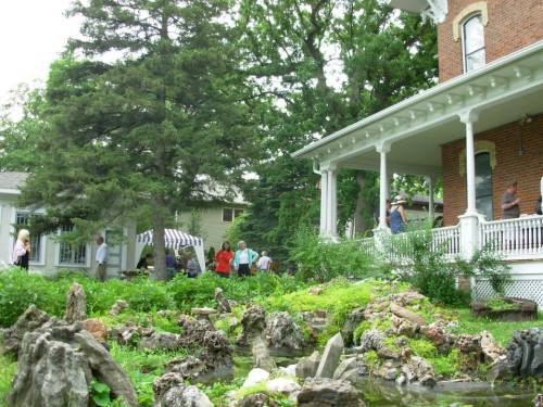A lush garden with rocky features and a historic house, people walking and enjoying the outdoor space.
