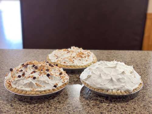 Three pies on a countertop, topped with whipped cream, one with chocolate chips and another with toasted coconut.