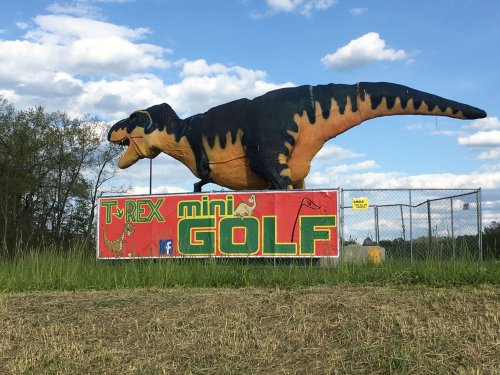 A large T-Rex statue atop a colorful sign for T-Rex Mini Golf, set against a blue sky with clouds.