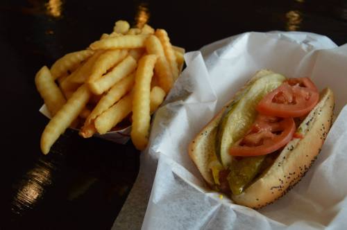 A hot dog topped with pickles and tomatoes, served with a side of crinkle-cut fries on a black table.