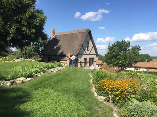 A thatched-roof house surrounded by lush green grass and colorful flowers, with people enjoying the garden.