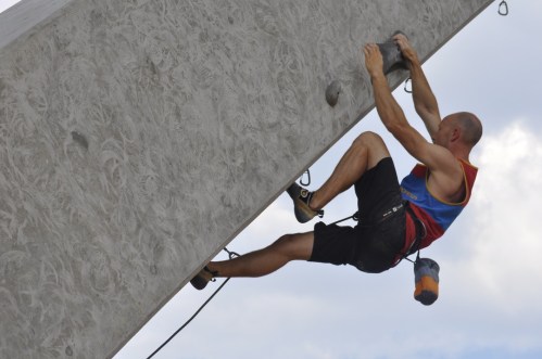 A climber ascends a vertical concrete wall, using handholds and wearing climbing gear.