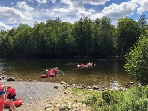 A sunny river scene with people in red rafts and a rocky shore surrounded by green trees and blue skies.
