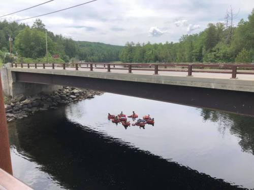 A bridge spans a calm river, with several red rafts floating below in the water, surrounded by green trees.