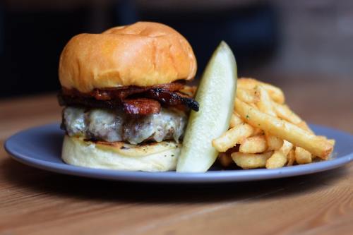A juicy burger with bacon on a bun, served with a pickle and a side of crispy fries on a gray plate.