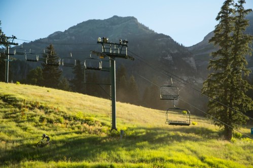 A cyclist rides through a grassy landscape with a ski lift and mountains in the background, illuminated by morning light.