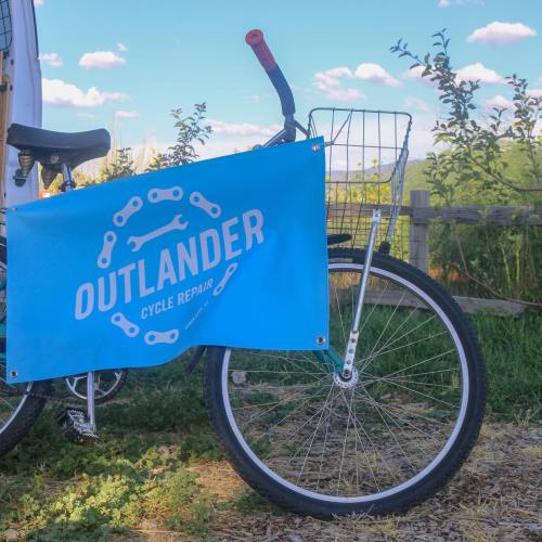 A blue banner reading "OUTLANDER CYCLE REPAIR" hangs on a bicycle with a basket, set against a scenic background.