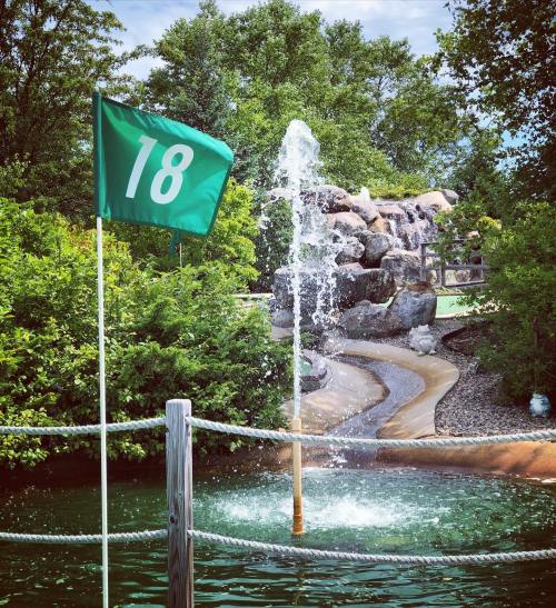 A green flag marked "18" near a water feature and rocky waterfall in a lush, landscaped area.
