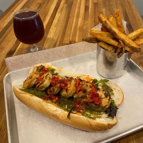 A shrimp sandwich topped with salsa on a tray, accompanied by a glass of dark beverage and a cup of fries.