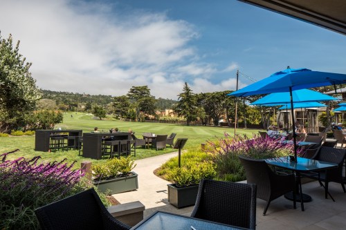 A scenic outdoor dining area with blue umbrellas, lush greenery, and a view of a golf course under a clear sky.