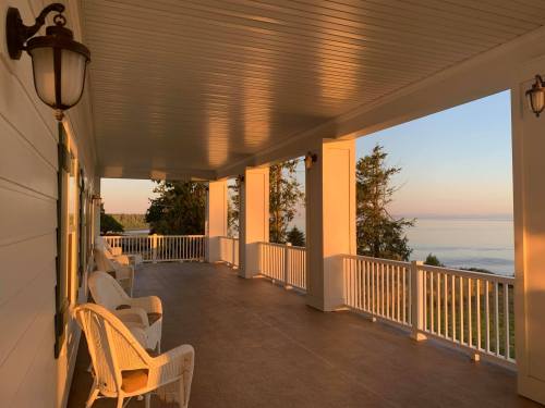 A serene porch with white chairs, overlooking a calm sea at sunrise, framed by tall trees and soft morning light.