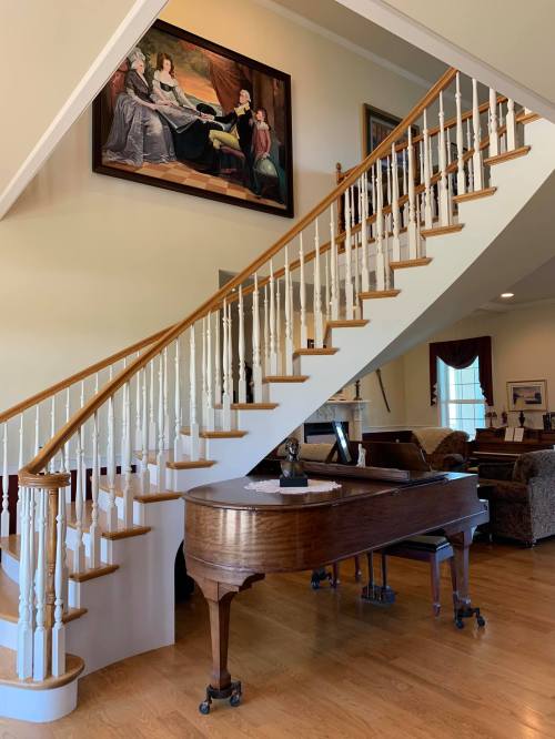 A wooden staircase curves upward beside a grand piano, with a large painting hanging on the wall above.