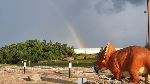 A large dinosaur statue in a parking area with a rainbow in the background and greenery on the hillside.