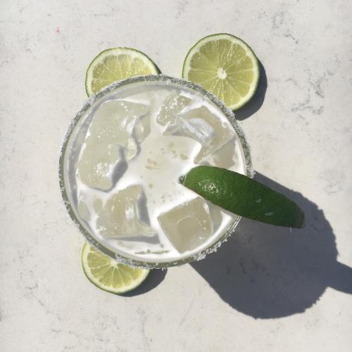 A refreshing cocktail with ice, garnished with a lime wedge, surrounded by lime slices on a light surface.