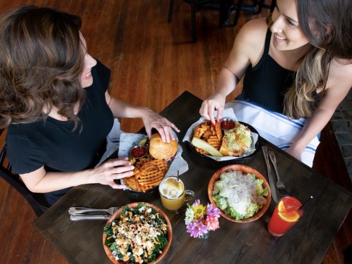 Two women enjoy a meal together, featuring burgers, fries, salads, and drinks on a wooden table.