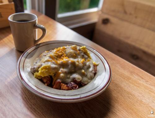 A plate of breakfast featuring scrambled eggs, gravy, and cheese over potatoes, with a cup of coffee beside it.