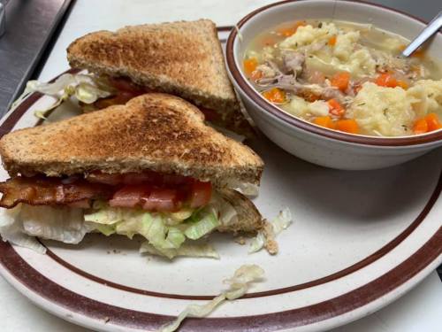 A plate with two toasted sandwiches filled with lettuce, tomato, and bacon, alongside a bowl of chicken and vegetable soup.