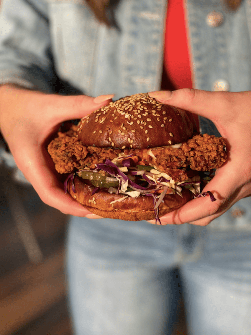 A person holds a sesame seed bun sandwich filled with fried chicken, pickles, and coleslaw, wearing a denim jacket.