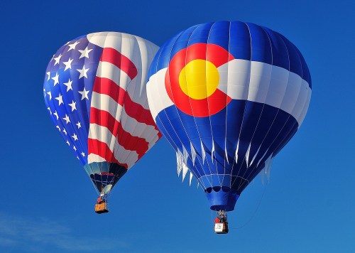 Two hot air balloons in the sky, one featuring the American flag and the other displaying the Colorado state flag.