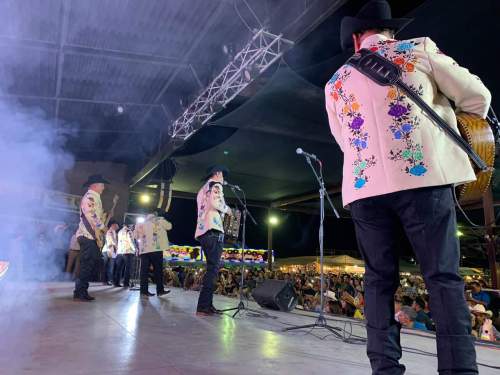 A group of musicians in colorful jackets perform on stage, with a crowd visible in the background and smoke effects.