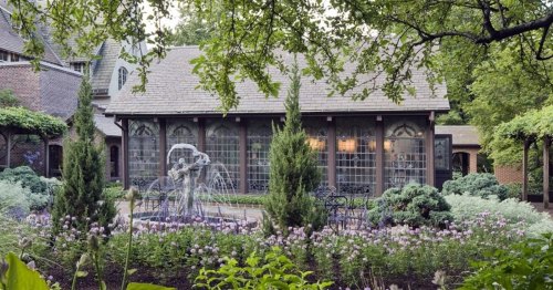 A charming garden with a fountain, surrounded by greenery and flowers, featuring a cozy building in the background.