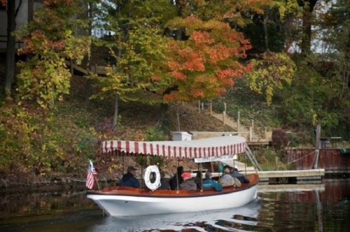 A boat with passengers glides through calm waters, surrounded by colorful autumn foliage and a wooden dock.